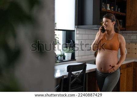 Beautiful pregnant young woman standing in the kitchen, speaking on the phone, touching her belly, smiling.