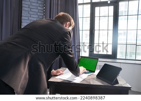 An unrecognizable businessman, he stands and types on his laptop using a chromakey on the background of the window. With a space to copy. High quality photo