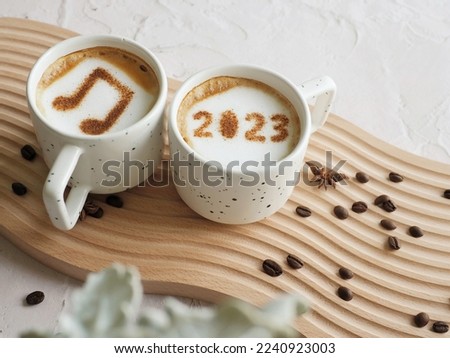 Coffee cups with number 2023 and musical note symbol over frothy surface flat lay on white cement background with coffee beans and star anise and plant. Happy new year 2023 food art theme music trend.