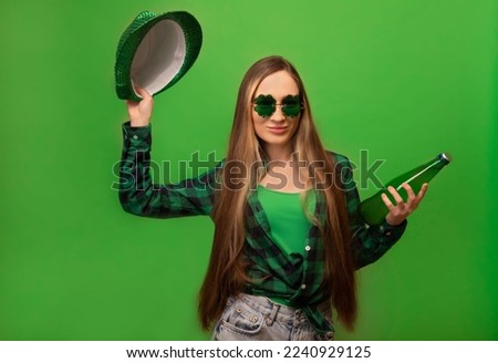 Happy smiling young woman in green checkered plaid shirt with clover shaped glasses holding green beer glass bottle and party hat isolated on green background.

St Patricks Day celebration concept.
