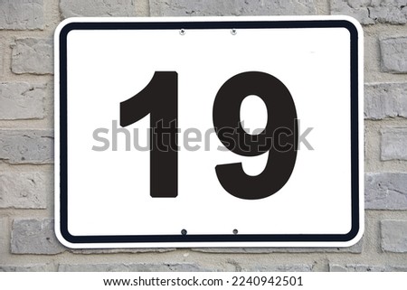 A white house number plaque, on a grey brickwall, showing the number nineteen (19) 