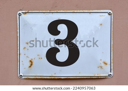 A white house number plaque, on a light red wall, showing the number three (3)
