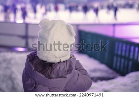 Back view portrait of a little girl standing by the ice rink and watching people.