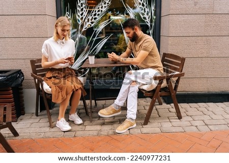 Side view of young couple using looking smartphones, ignoring each other sitting at table with coffee in street cafe on summer day. Concept of internet more interesting than real communication. Royalty-Free Stock Photo #2240977231