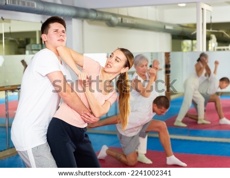 Concentrated young woman paired with male partner in self defense training, practicing basic elbow kick to chin during back grab