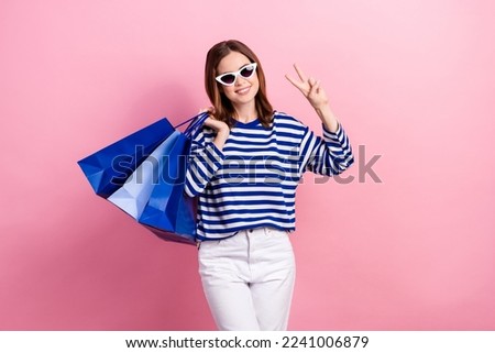 Photo of pretty positive girl hold boutique bags hand fingers show v-sign isolated on pink color background