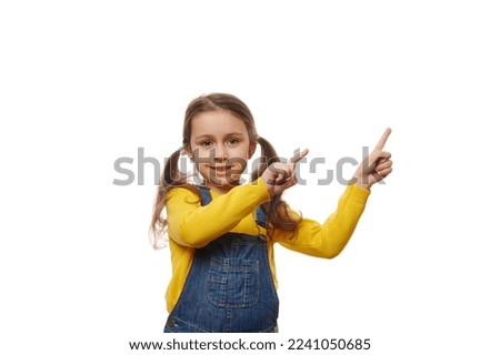 Emotional studio portrait of a charming little girl with two ponytails, wearing bright yellow sweater and denim overalls, smiling at camera and pointing fingers at copy ad space for promotional text.