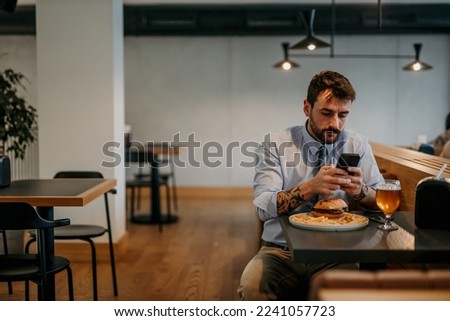 Focused businessman sitting alone in the local cafe, having a lunch break with a burger and beer and texting on his phone. Royalty-Free Stock Photo #2241057723