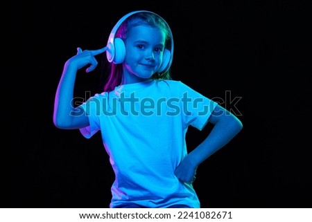 Portrait of little beautiful girl, child in white T-shirt posing in headphones isolated over black studio background in neon light. Concept of childhood, emotions, facial expression, lifestyle