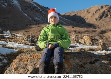 Portrait of handsome cute boy sitting against the background of the mountains dressing Santa hat and looks at the camera. new year 2023 in the mountains. Beautiful clear blue sky
