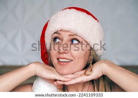 Smiling Santa girl wearing red hat is laying her chin on both hands looking up side. Portrait of beautiful young blue-eyed blonde woman in Santa's hat. Christmas portrait. Christmas holidays, vacation