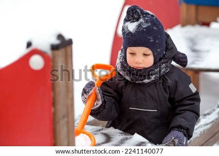 A little boy walks on the playground. A child cleans snow with a child's shovel. Childhood and teaching children to work. Snow removal after a snowfall.