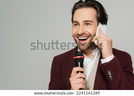 cheerful showman talking on smartphone while holding microphone isolated on grey