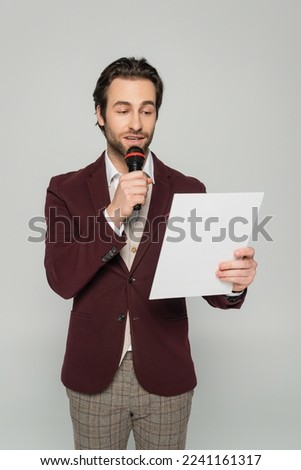 bearded host of event in formal wear making announcement in microphone while reading from paper isolated on grey