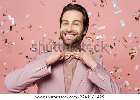 Smiling host of event adjusting bow tie near blurred confetti on pink background