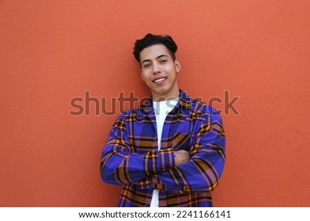 Young man of hispanic heritage wearing colorful flannel shirt standing with his arms crossed near the orange wall. Copy space for text, close up, background.