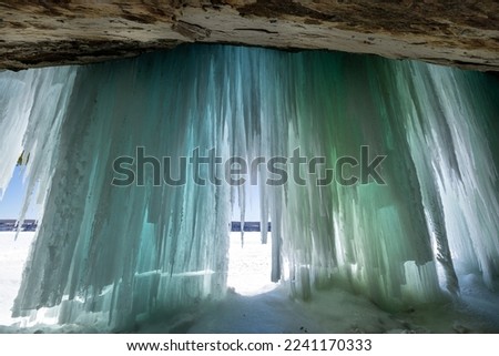 Blue and green ice curtains drape over the entrance of a Lake Superior ice cave on Grand Island during winter. Long shadows from the rising sun cross the ice floor of the cavern.
