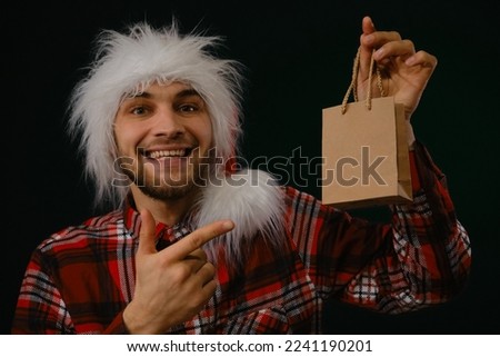 Young Caucasian man in red fluffy Santa Claus hat holds eco friendly craft package in hands and gives gift. Studio portrait on dark background. Concept Christmas promotion sale on Black Friday.
