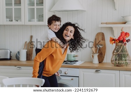 Cheerful son at kitchen riding  on moms back laughs happy spend time together. Curly Spanish woman I orange sweater entertains little boy home. Playful Italian kid hugs babysitter. Happy family. Royalty-Free Stock Photo #2241192679