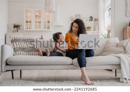 Overloaded  young Italian woman with curly hair dressed in orange blouse and dark blue pants sitting on sofa using laptop, working at home at the kitchen with son playing on phone, remote working. Royalty-Free Stock Photo #2241192685