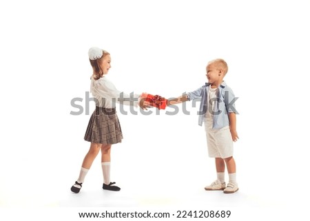 Portrait of beautiful little children, boy giving present box to girl isolated over white studio background. Concept of childhood, friendship, game, school, fun, education. Copy space for ad
