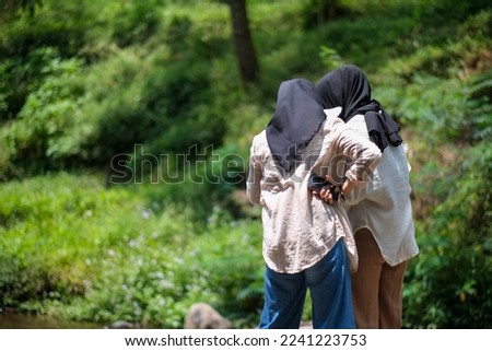 Two young Asian women, wearing hijabs, stand close together and back to back. Green hill background.