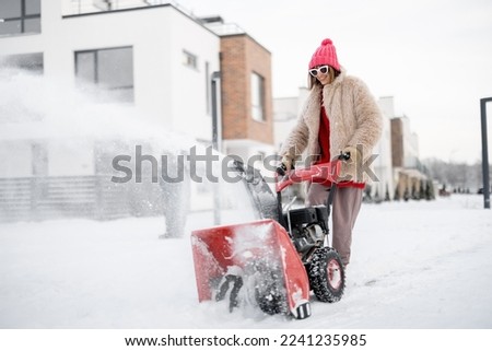 Woman removes snow with a snow thrower machine near house at residential area. Winter yard care and easy technology concept Royalty-Free Stock Photo #2241235985