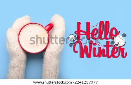 Hands in knitted mittens and with cup of tasty cacao drink on blue background with text HELLO WINTER