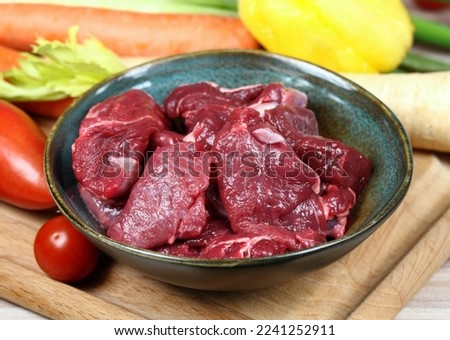 Raw deer meat for venison ragout or goulash. Bowl with pieces of deer meat on cutting board, fresh vegetable for cooking around. Royalty-Free Stock Photo #2241252911