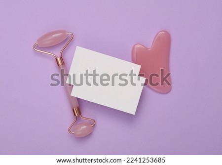 White Blank business card for branding and jade facial massage roller and gua sha scraper on purple background. Beauty concept. Top view
