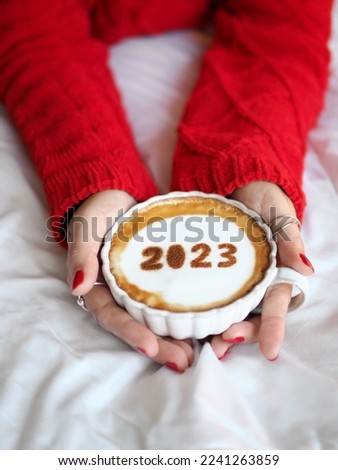 Happy New Year 2023 holidays food art theme number 2023 over frothy surface of cappuccino served in coffee cup holding by woman in red knitted sweater lying on bed with white blanket. New year new you
