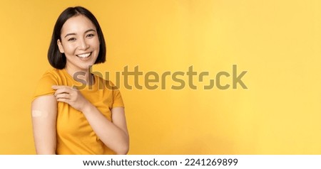 Vaccination and covid-19 pandemic concept. Happy and healthy asian girl pointing at her shoulder with band aid after vaccinating from coronavirus, yellow background.
