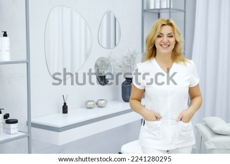 Beautiful mature woman cosmetologist-aesthetician in a medical suit posing against the backdrop of her office. Horizontal photo Royalty-Free Stock Photo #2241280295