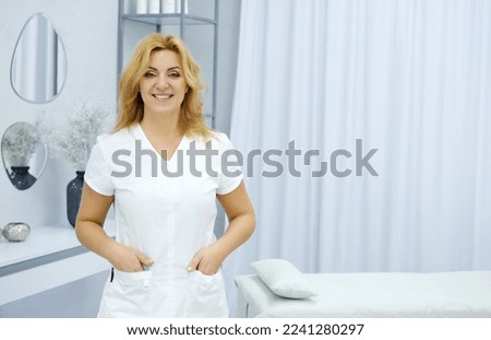 Beautiful mature woman massage therapist in a medical suit posing against the backdrop of her office. Horizontal photo