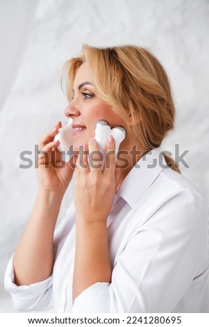 Mature blonde Caucasian woman with a natural make-up and healthy fresh glowing skin using the electric facial massager. Home skin care concept. Vertical photo