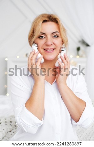 Mature blonde Caucasian woman with a natural make-up and healthy fresh glowing skin using the electric facial massager. Woman is looking at camera and smilling. Vertical photo