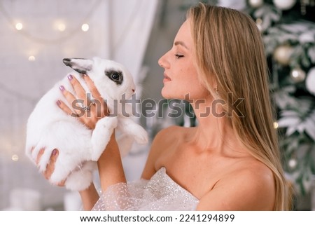Woman holding a white rabbit symbol of the year 2023. Close-up of a beautiful young blonde woman holding a rabbit in a sparkly dress. She sits in a Christmas decorated room