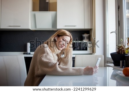 beautiful young woman in a cozy sweater sits in the kitchen and watches updates on social networks. Girl at home with phone in hand