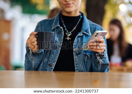 Woman sitting outdoors cafe drinking beverage holding in hand smartphone