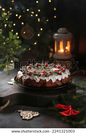 A Christmas cake decorated with white icing, cranberries and rosemary stands on a wooden board, against a dark background, in the background a lantern and bokeh