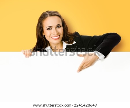 Happy smiling brunette young business woman in blue clothing, standing behind, peeping from blank banner or mock up signboard, showing copy space for text, on yellow background.