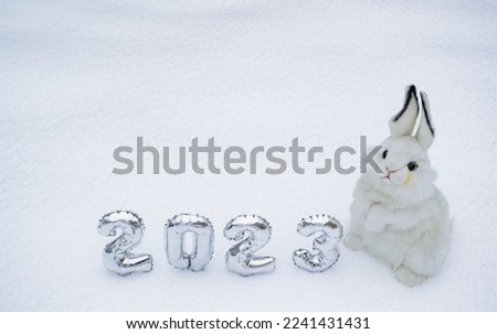 White fluffy bunny toy with silver numbers of the New Year 2023 on the white snow background. Copy space. Symbol of Chinese New Year 2023 