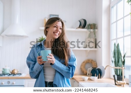 Happy caucasian female with long loose hair in jeans and blue shirt toothy smiles holds cup of tea looks aside happily enjoys weekend at cozy home. Pretty hispanic woman laughs against blurry kitchen. Royalty-Free Stock Photo #2241468245