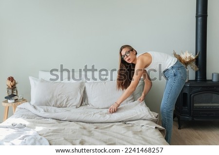 Confident caucasian beautiful young woman makes bed wears jeans and white t-shirt and glasses. Pretty hispanic girl makes housekeeping. Mockup, people at home. Young  student makes order home.