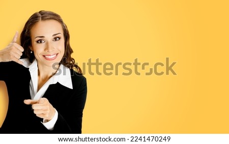 Businesswoman in black confident suit showing call me hand sign gesture and pointing at viewer, on yellow background. Portrait of smiling brunette woman at studio. Business concept.