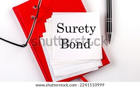SURETY BOND text on a sticker on red notebook with pen and glasses Royalty-Free Stock Photo #2241510999