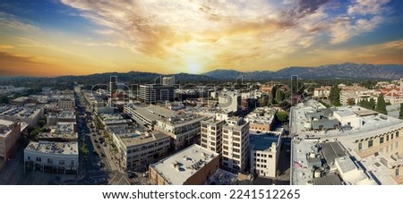 an aerial shot of the apartments, shops and office buildings in the city skyline with mountains and cars driving on the street surrounded by lush green trees and powerful clouds at sunset in Pasadena Royalty-Free Stock Photo #2241512265
