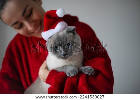 Smiling happy woman hugs her cute Scotland shorthair cat in Santa Christmas hat. Having fun with pet at home. Christmas and New year concept