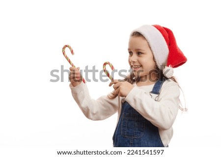 Caucasian 5-6 years child, cute baby girl in Santa hat, pointing with green red striped candy canes at copy advertising space for Christmas, on white background, expressing happiness and cheerfulness
