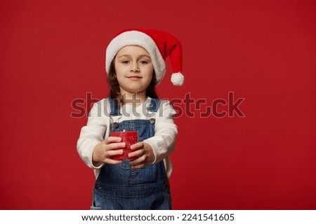 Caucasian lovely child, adorable blue-eyed little girl wearing Santa hat and blue denim overalls, cutely smiling looking at camera, holding out a lit Christmas candle, isolated on red background